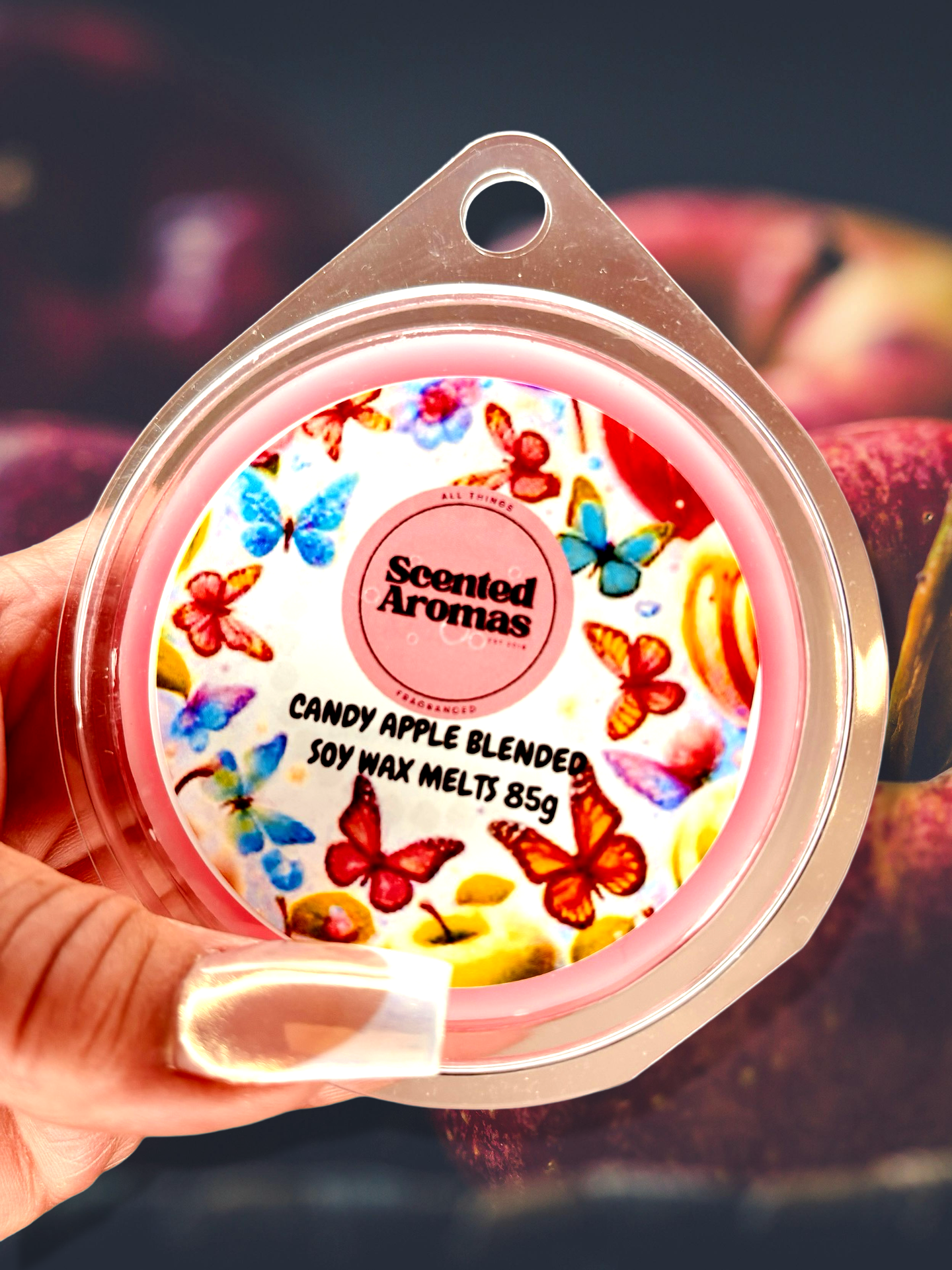 A hand holds a round package labeled "Scented Aromas," adorned with butterflies, featuring the text "Candy Apple Wax Melts 85g." Discover high-quality wax melts in the UK, meticulously crafted for a delightful aromatic experience.