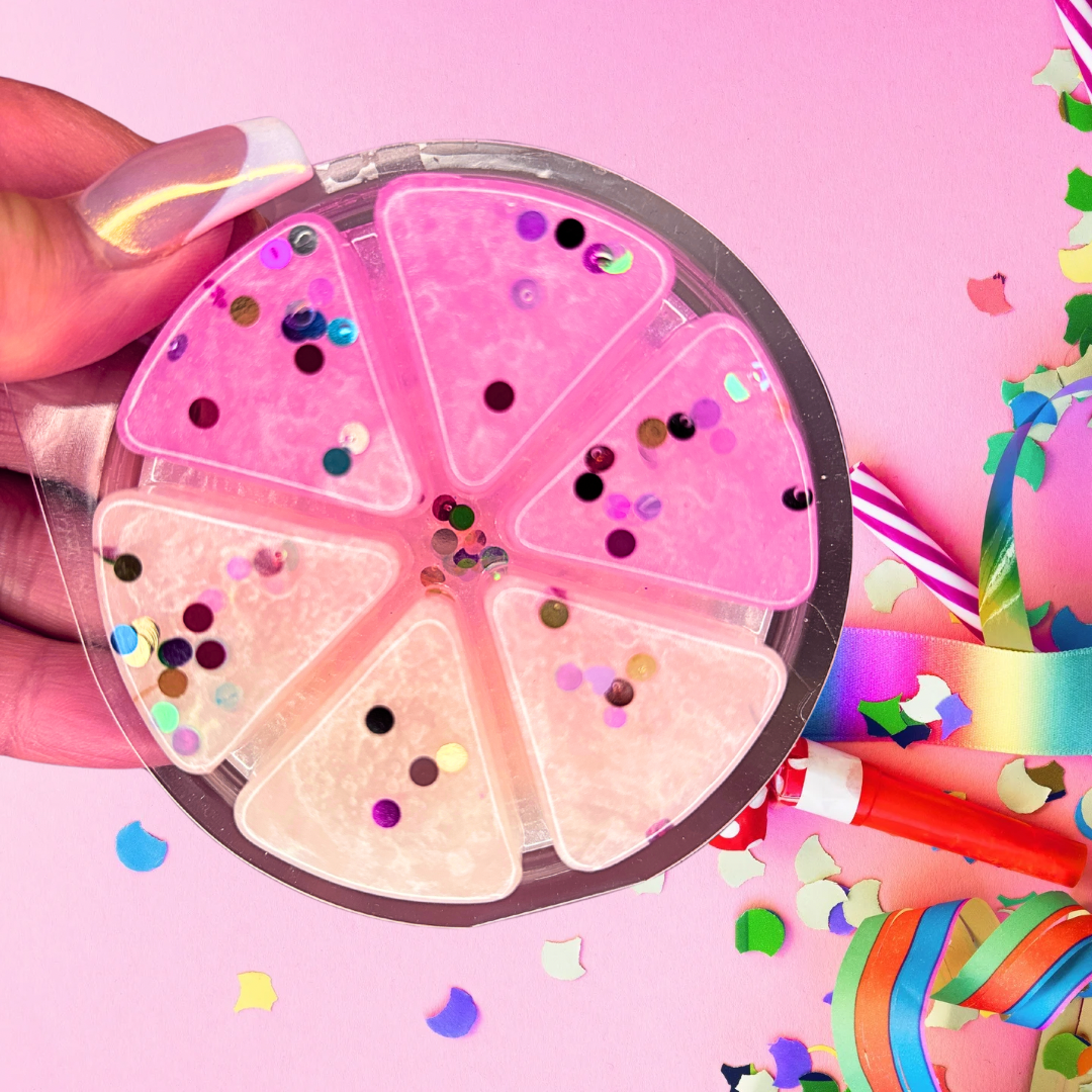 A hand holds a round container of Carnival Treats Gel Wax Melts by The Soap Gal x, featuring six compartments. Set against a pink background, it's adorned with confetti-like colorful paper shapes and twisted ribbons.