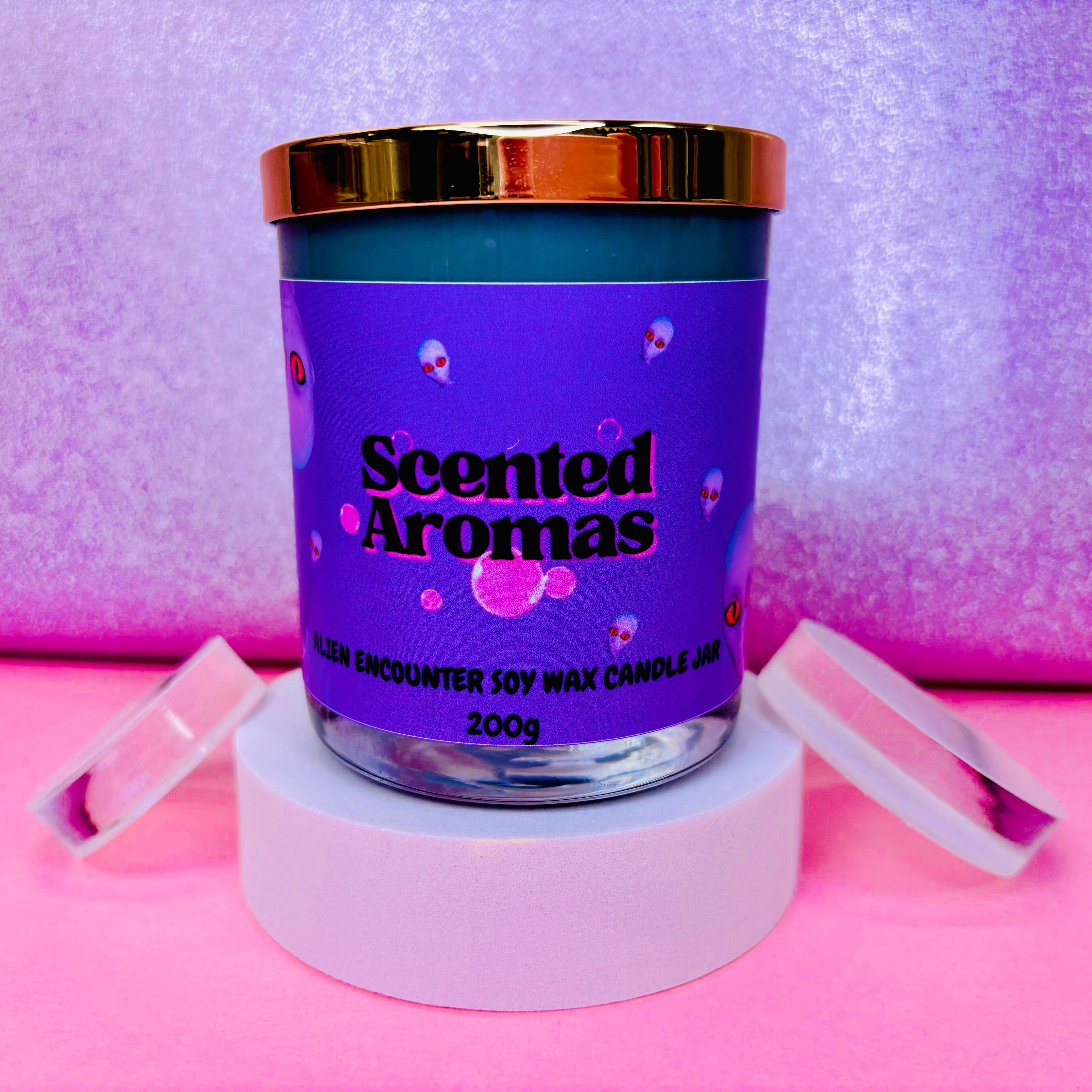 The Alien Encounter Candle - 200g by Scented Aromas rests elegantly on a white stand, casting a soy wax glow against purple and pink hues, adding coziness to any home.