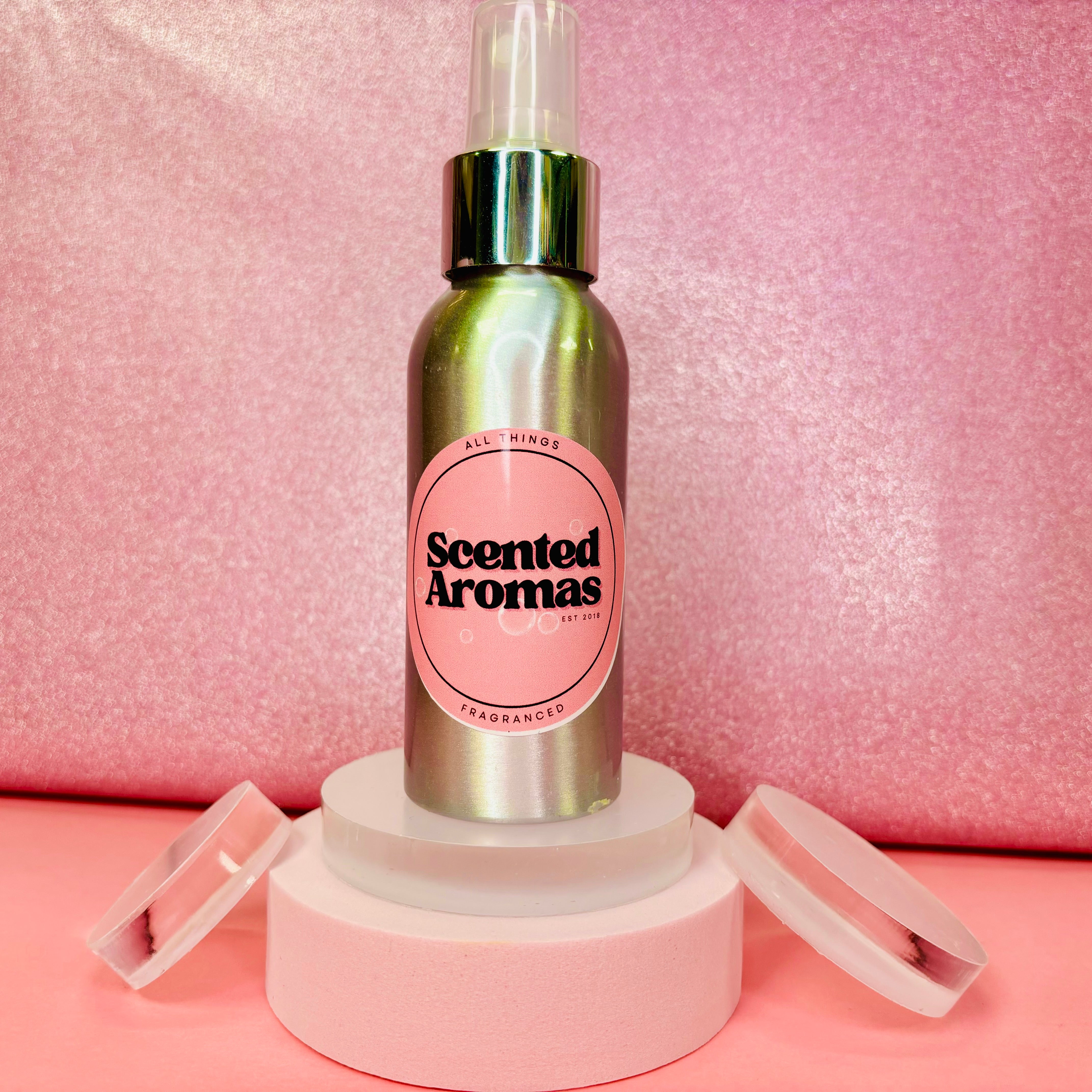 A metallic bottle of Scented Aromas' Rouge Inspired Body Spray stands on a white display with two open lids beside it, set against a pink background.