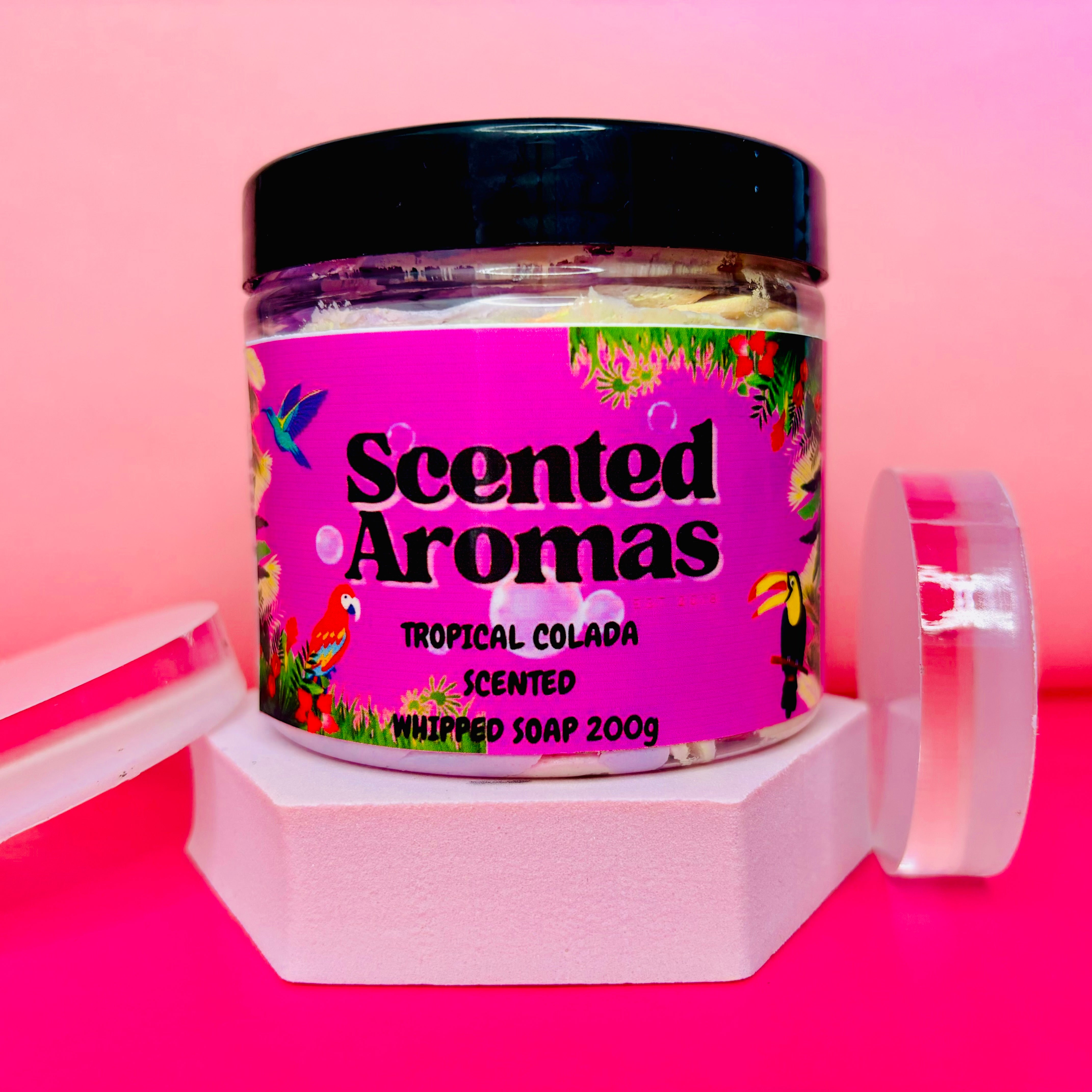 A 200g jar of "Unicorn Fluff Tropical Colada" whipped soap from The Soap Gal x, with a pink label and black lid, is stylishly presented on a white hexagonal stand against a pink backdrop.