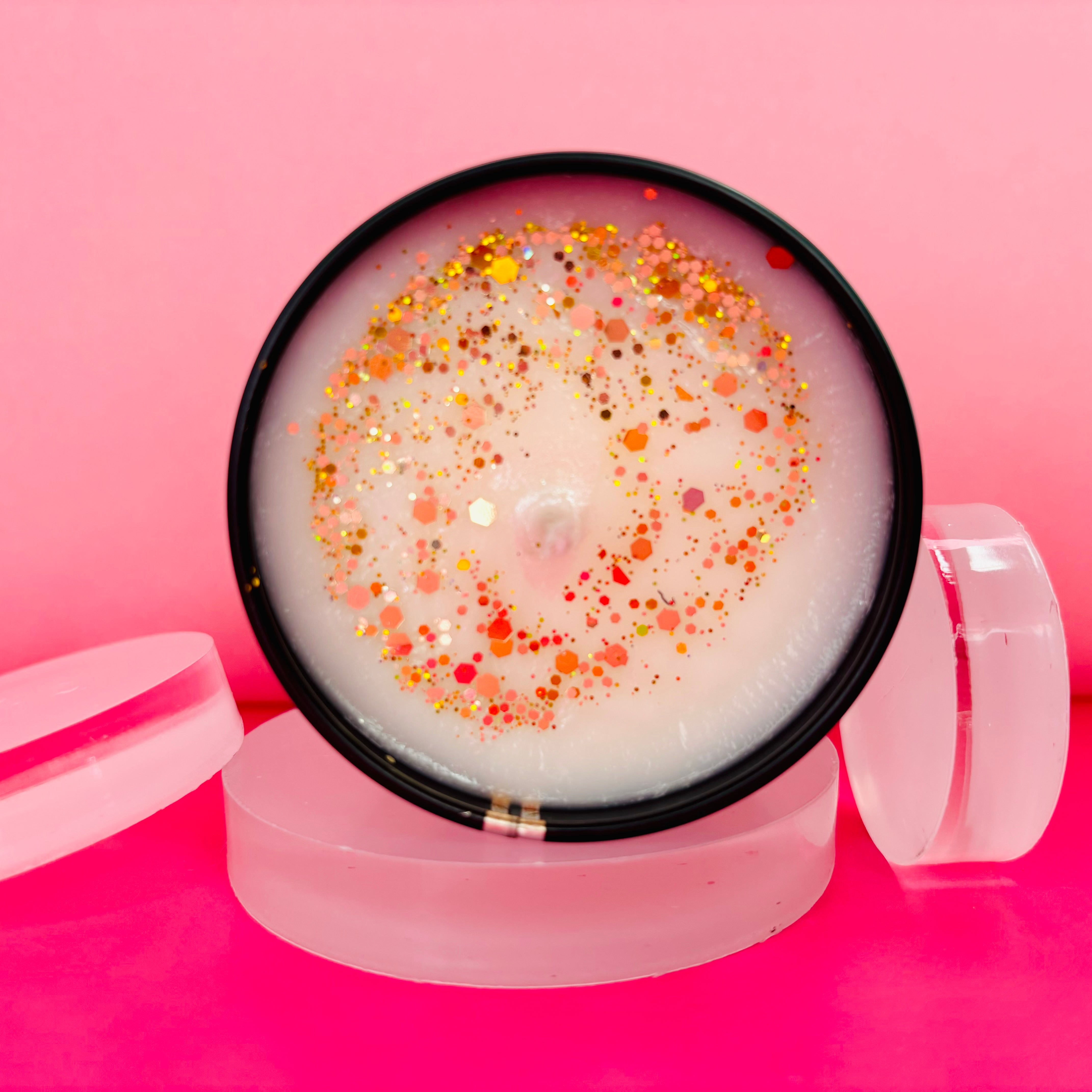 Introducing the Sticky Toffee Pudding Candle Tin - 100ml by The Soap Gal x, a round candle adorned with gold and orange glitter, beautifully displayed on a clear stand against a pink background. Created from soy vegan-friendly wax, this candle ensures long-lasting fragrances to enhance your space.