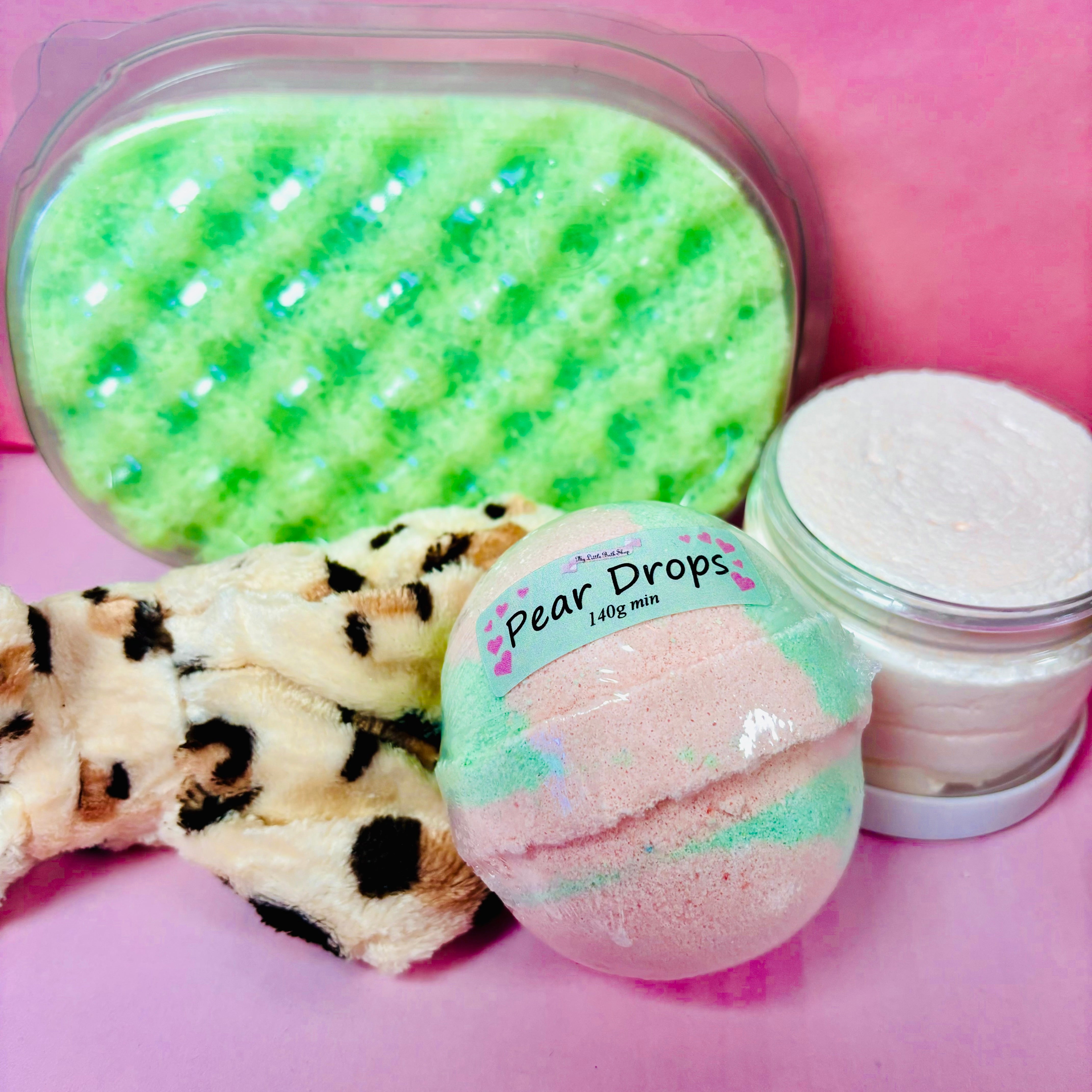 A Scented Aromas set with a "Pear Drops" bath bomb, leopard print headband, green soap sponge, and body scrub against a pink background evokes delightful scented aromas.