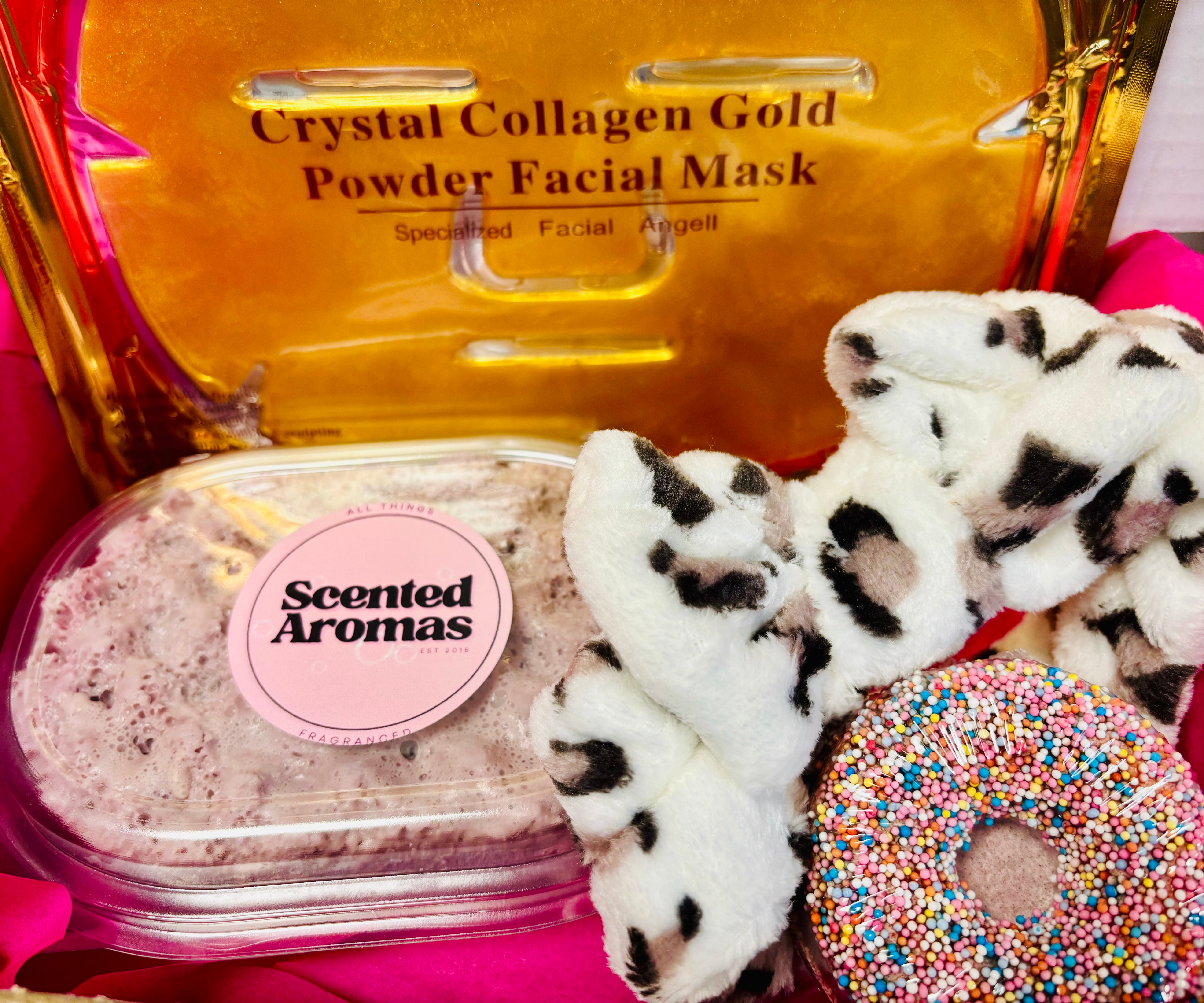 The Scented Aromas Girls Night In Self Care Bath and Body Set includes a soap sponge, bath bomb, collagen face mask, and headband. Perfect for your self-care routine, it's all presented beautifully with a colorful doughnut against a pink background.