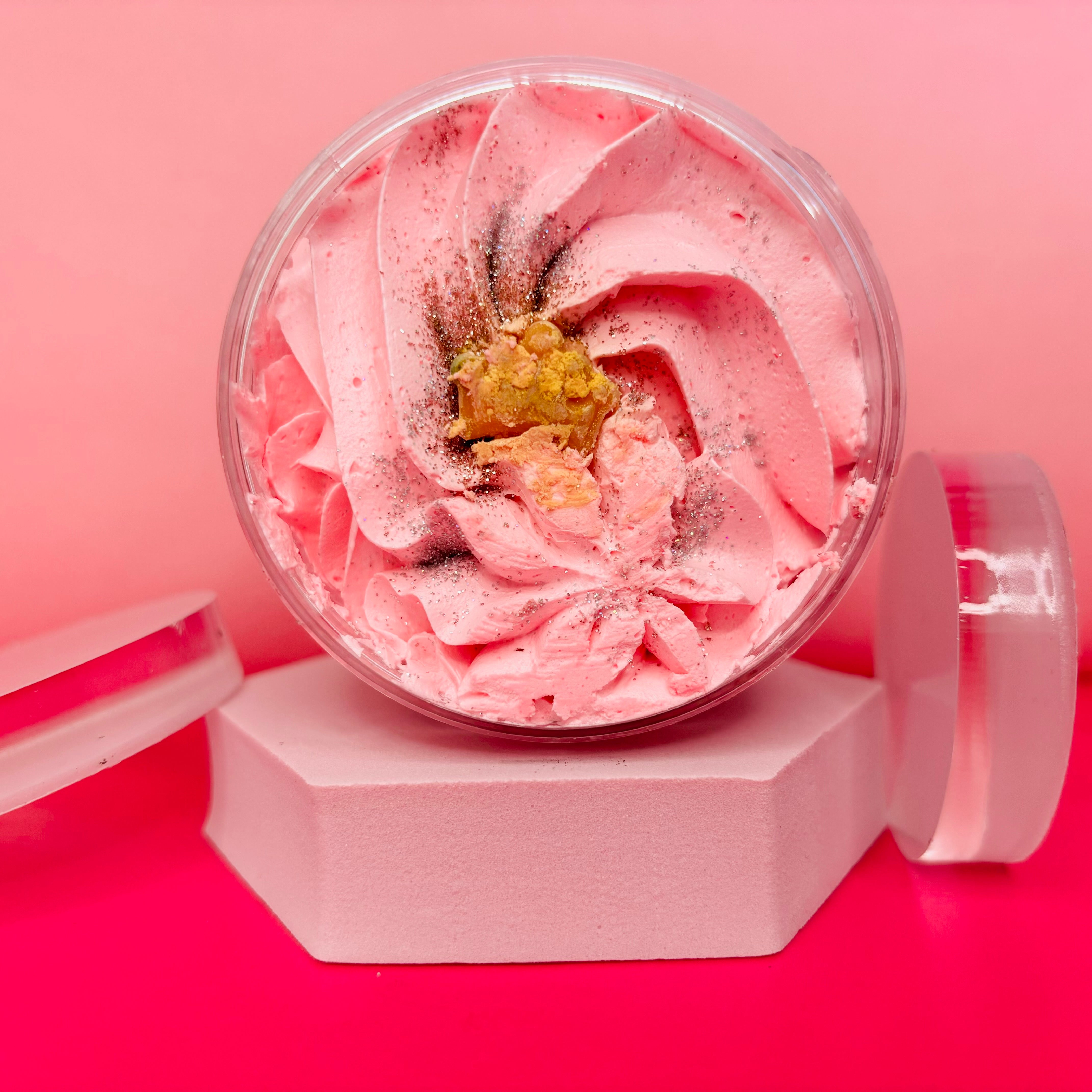 A jar of Belle Of The Ball Whipped Soap Shave Butter, adorned with a sprinkle of glitter and a small piece of gold leaf on top, reminiscent of a La Vie Est-inspired perfume by The Soap Gal x, placed elegantly on a white hexagonal stand.