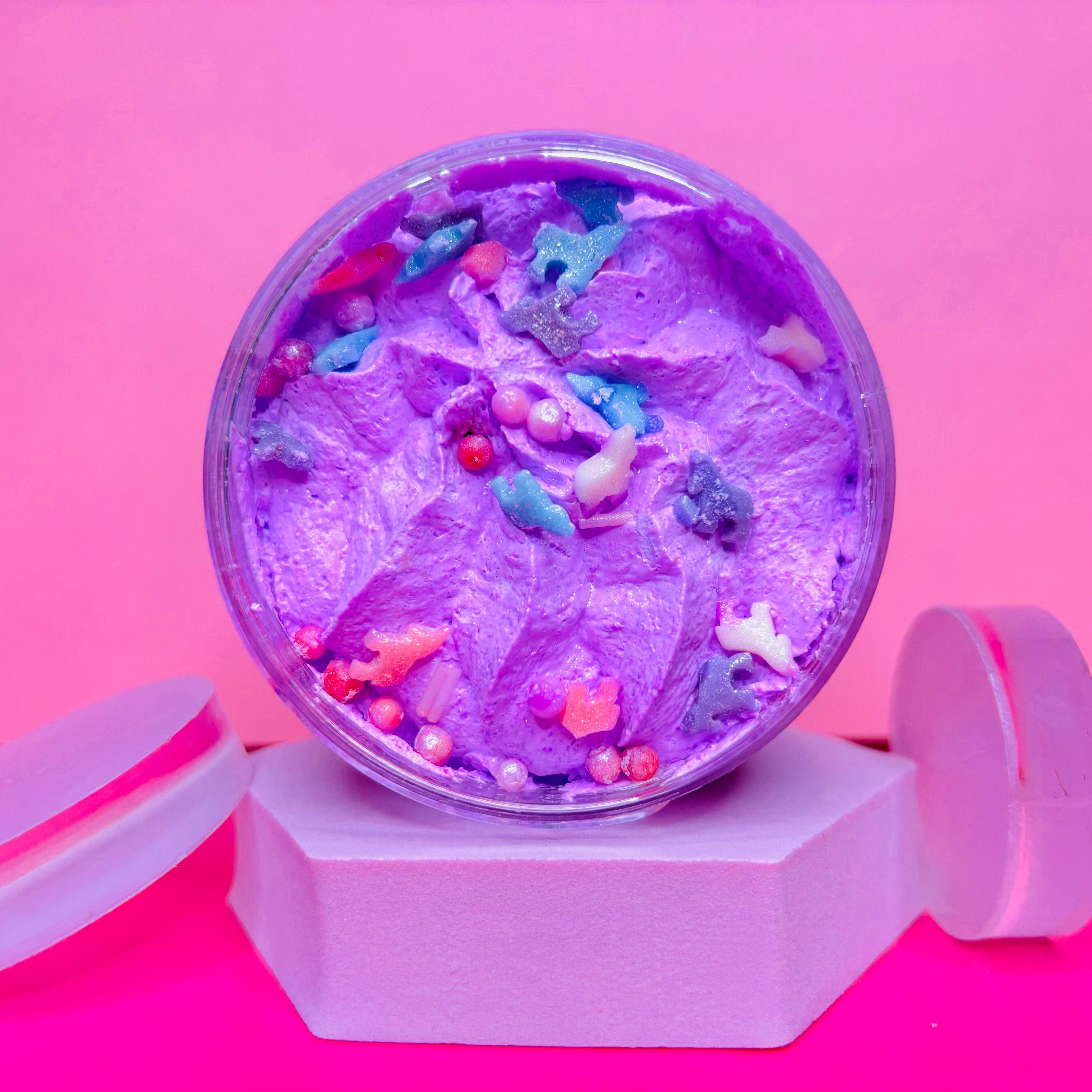 A jar of purple whipped body butter with colorful star and bead decorations sits on a white hexagonal platform against a pink background, evoking the charm of Scented Aromas' Birthday Cake scented Bath Bomb. Ideal for any skincare gift set or part of the Mother's Day Gift Set Bundle.