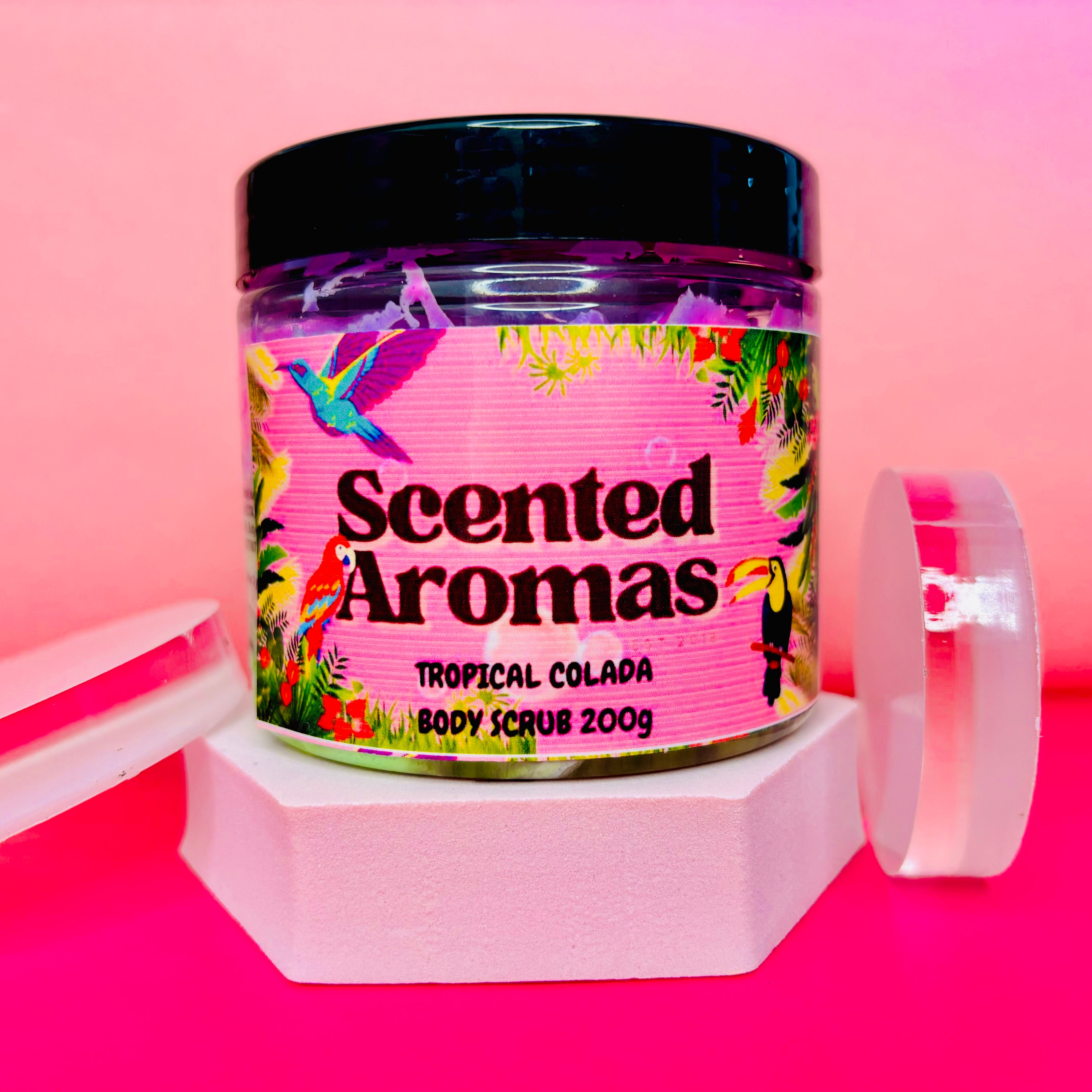 A 200g jar of The Soap Gal x Unicorn Fluff Tropical Colada Scented Body Scrub sits against a vibrant pink and red backdrop. Its tropical-themed label promises an exotic experience as it gently exfoliates your skin.