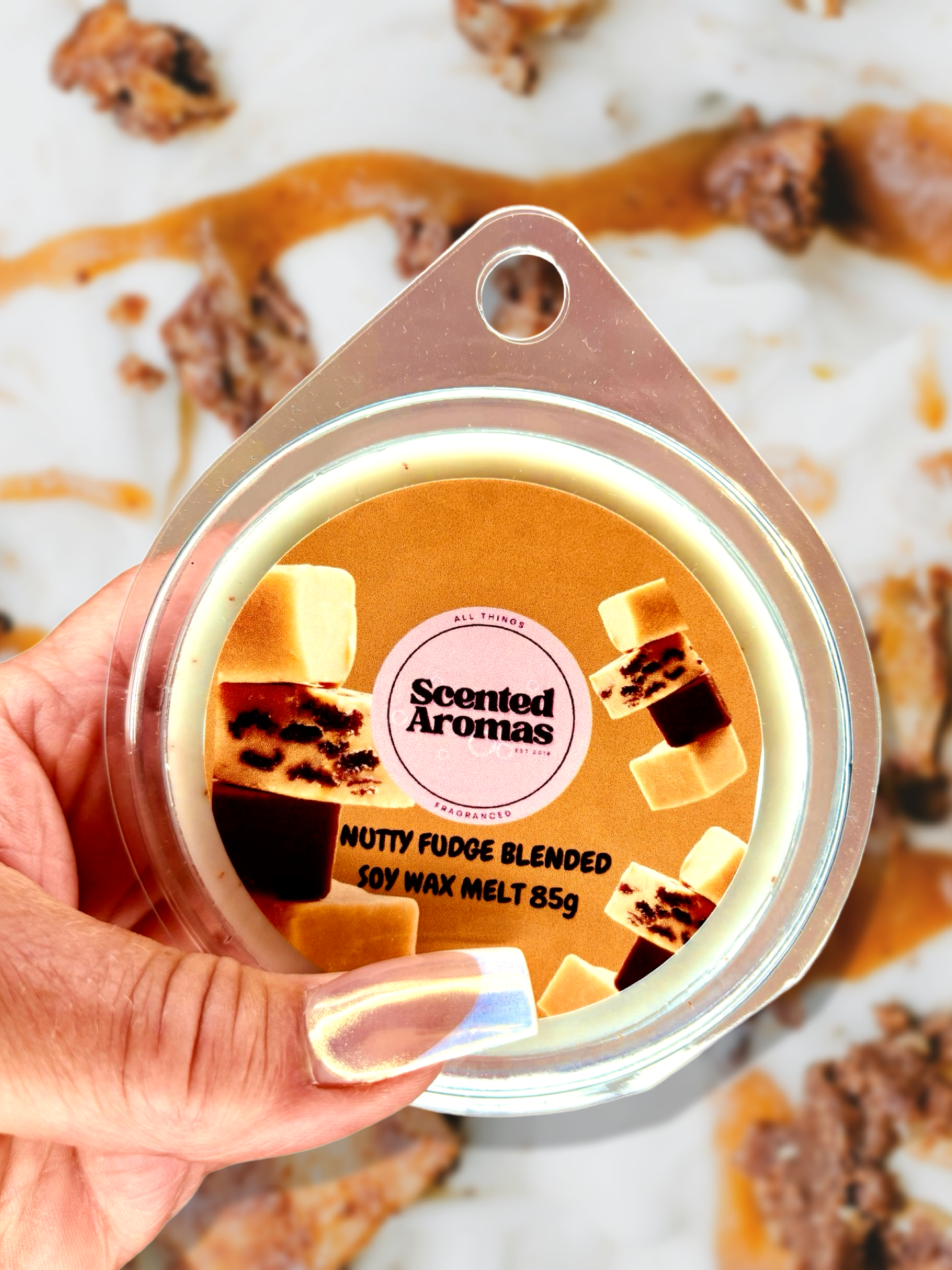 A hand holds a container of "Nutty Fudge Wax Melt 85g" by The Soap Gal x, enriched with a long-lasting fragrance. The background features ingredients such as nuts and caramel, emphasizing the enchanting aroma of the soy wax blend.