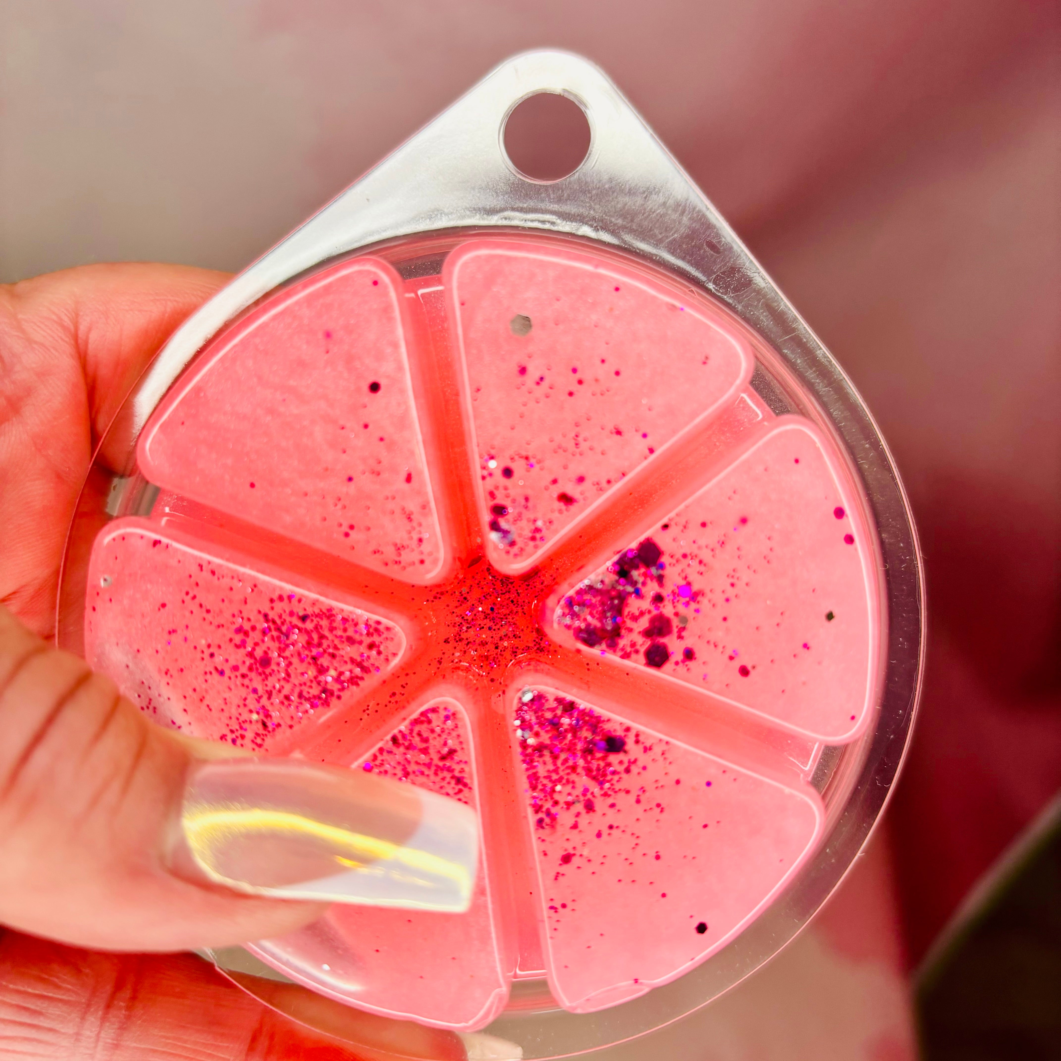Close-up of a person holding a clear plastic container with The Soap Gal x's Le Est Belle Gel Wax Melts, 85g, featuring pink, segmented luxury wax melts and scattered magenta specks inside.