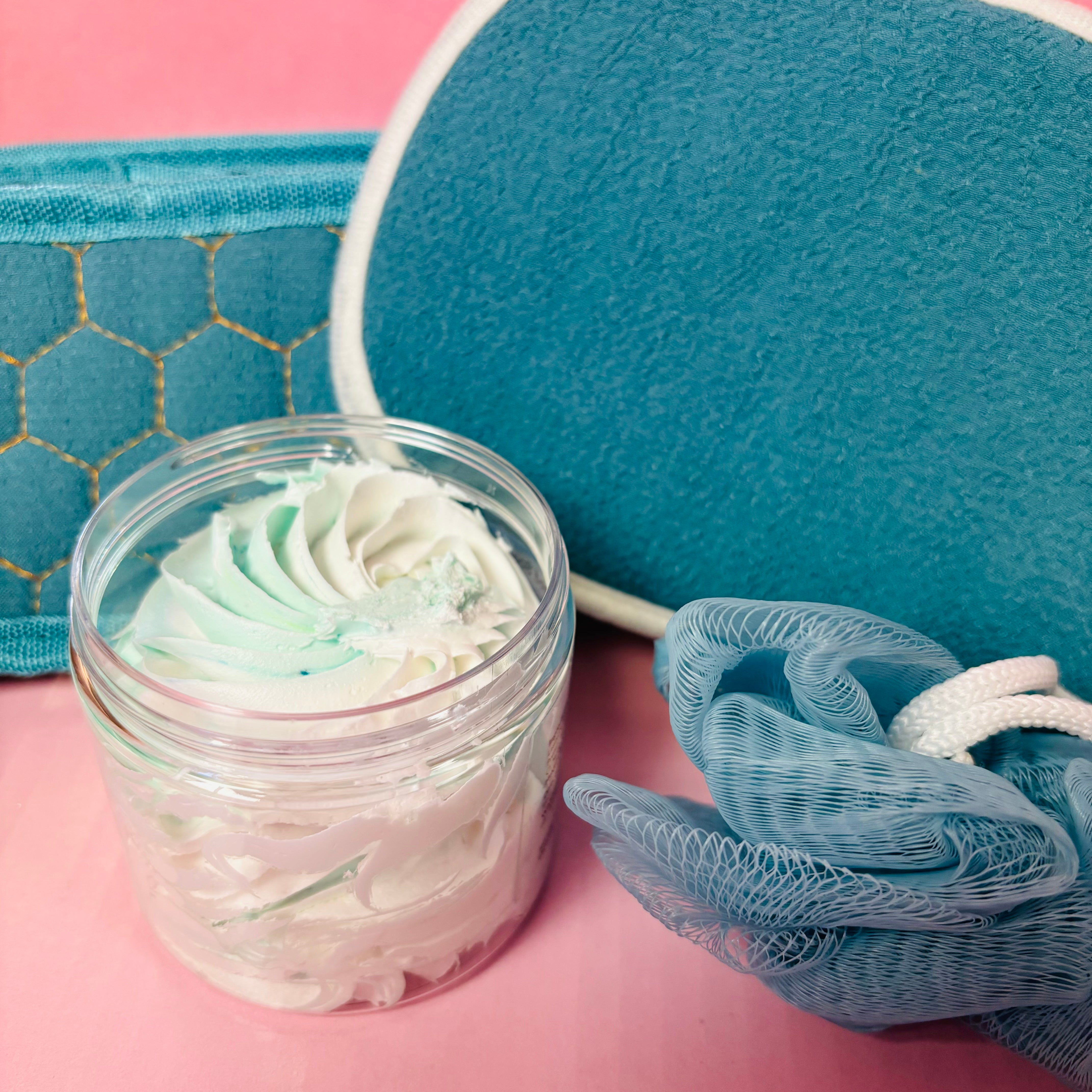 The Scented Aromas 4 Piece Bath and Body Gift Set, featuring whipped soap, a blue loofah, a teal honeycomb-patterned band, and a round cushion on a pink surface, offers soothing textures and aromas perfect for enhancing your self-care routine.