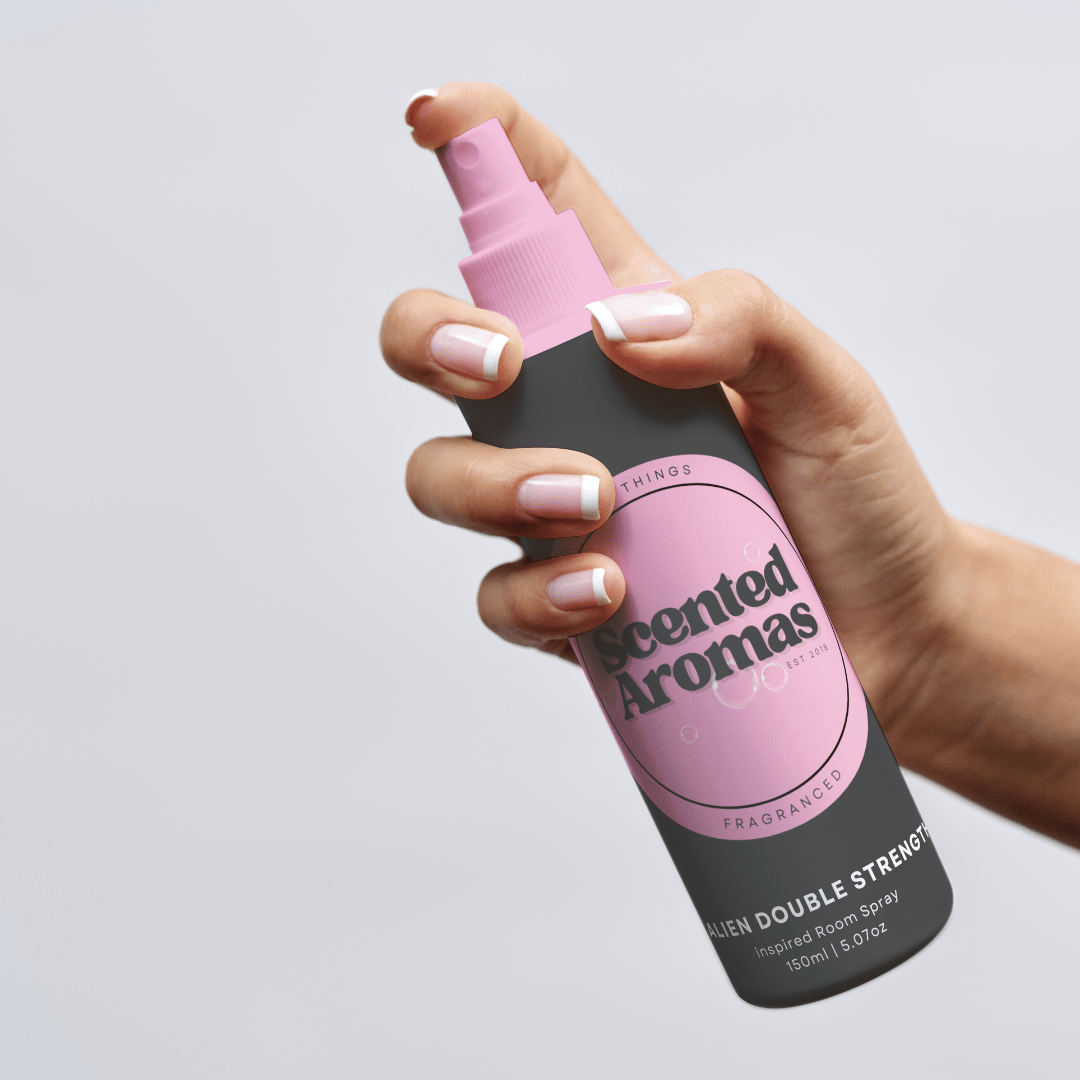 A hand holding a bottle of The Soap Gal x Alien Encounter Room Spray 150ml, adorned with a pink label, unveils its enchanting woody floral blend, designed as an effective odor eliminator for any space.