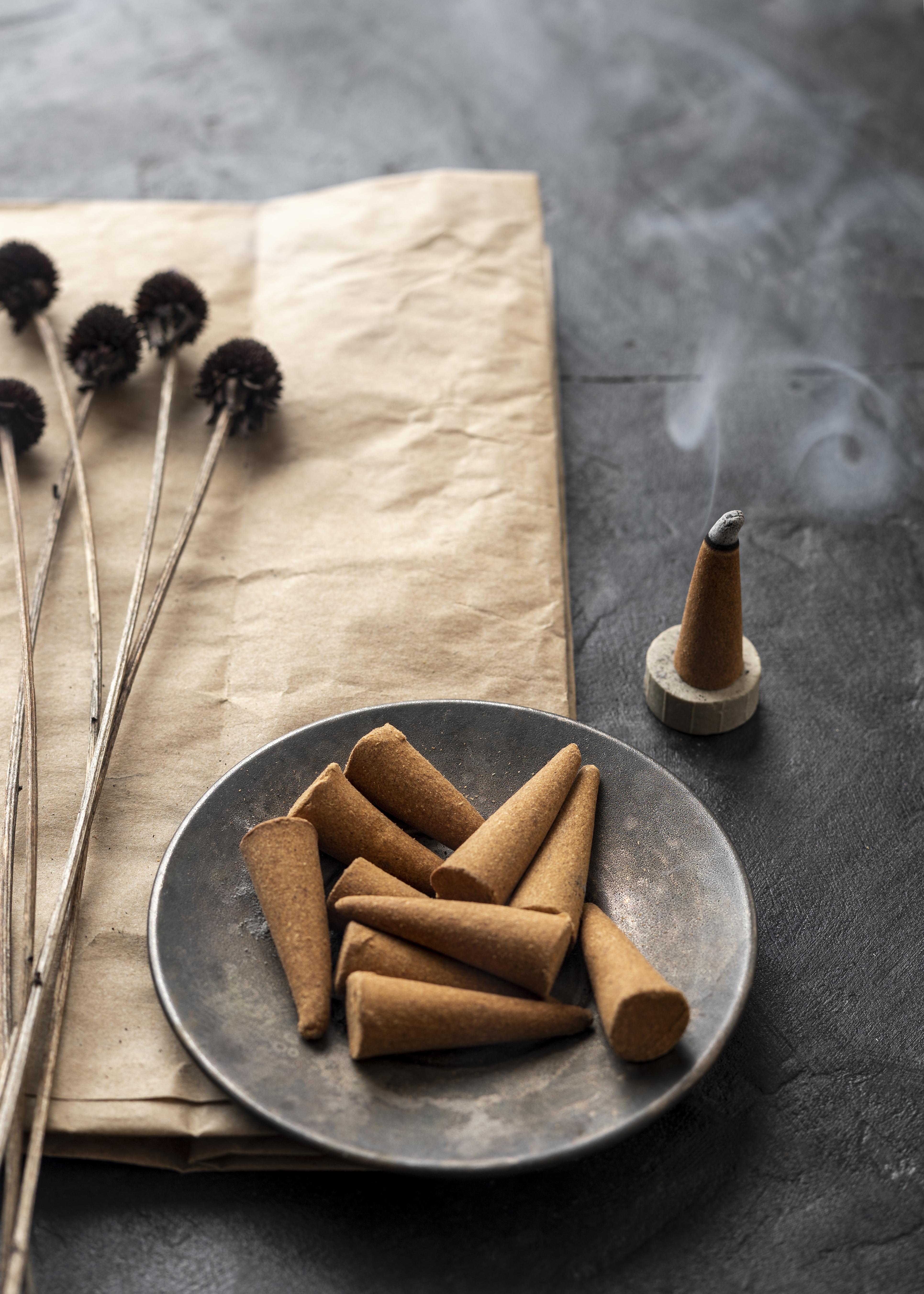 Palo Santo Incense Cones from The Soap Gal x sit on a round plate, with smoke gracefully rising from an ignited cone nearby, evoking the essence of spiritual rituals. In the background, dried Palo Santo flowers lie on brown paper, fostering an aura of calm and reflection.