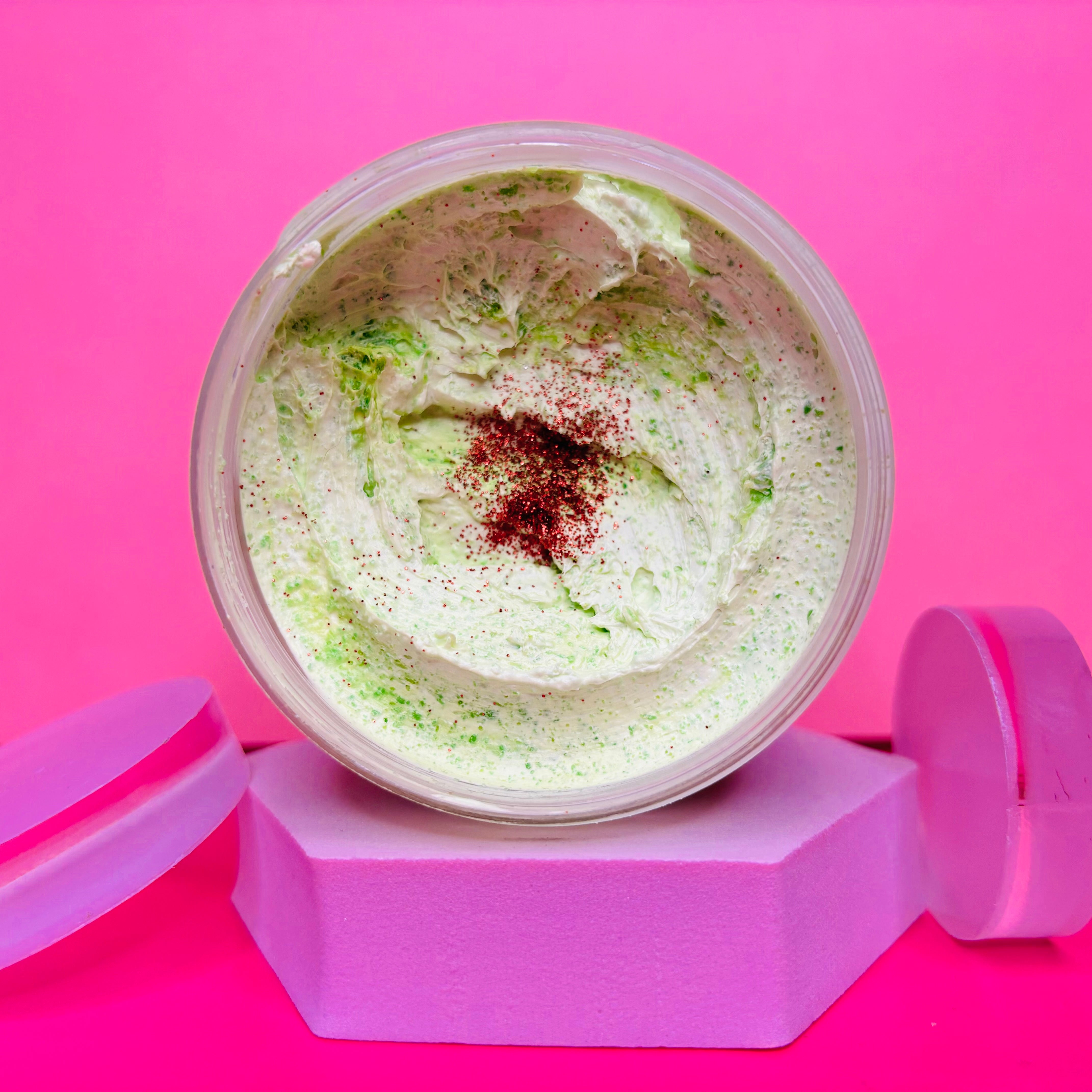 An open jar of Cinnamon and Apple Sugar Body Scrub 200g from The Soap Gal x, featuring a creamy texture with green flecks and topped with pinkish-red powder, rests on a pink octagonal stand against a vibrant pink backdrop. This scrub is crafted to enhance skin circulation while delighting your senses.