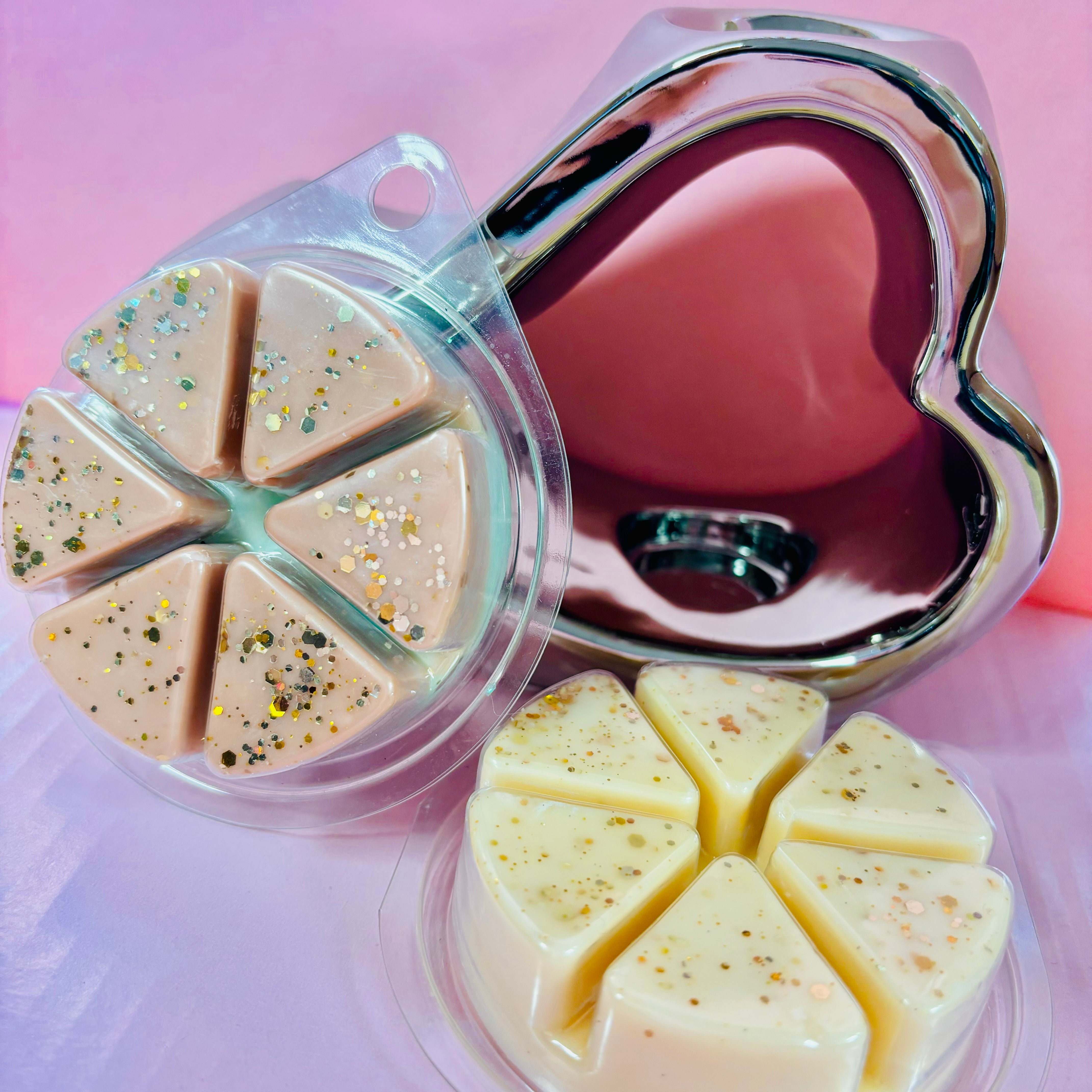 A pink and cream heart-shaped wax melt set with gold glitter, accompanied by a silver heart ornament against a pink backdrop, forms the Wax Warmer Best Seller from Scented Aromas—a charming addition to any collection.