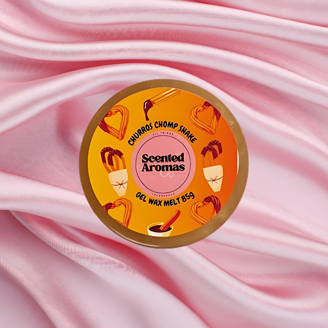 A round container labeled "Scented Aromas," featuring chocolate and caramel designs, sits on pink satin fabric. The Soap Gal x's Fresh Churros Magical Gel Wax Melts, 85g, offer a long-lasting indulgent fragrance experience reminiscent of churros.
