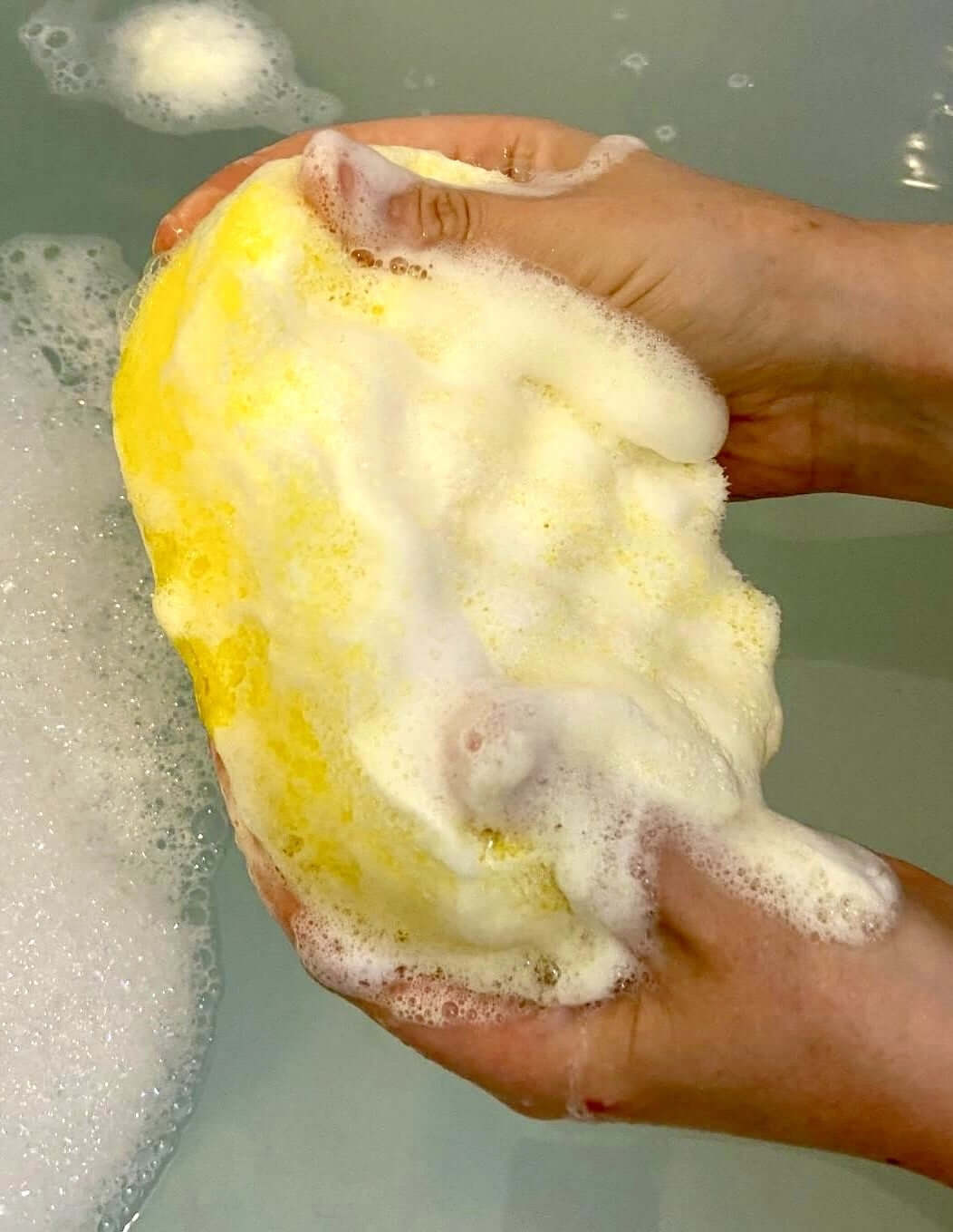 Hands scrubbing The Soap Gals' Citronella Soap Sponge - Natural Insect Repellent with rich soap lather, next to a bubbly water surface.