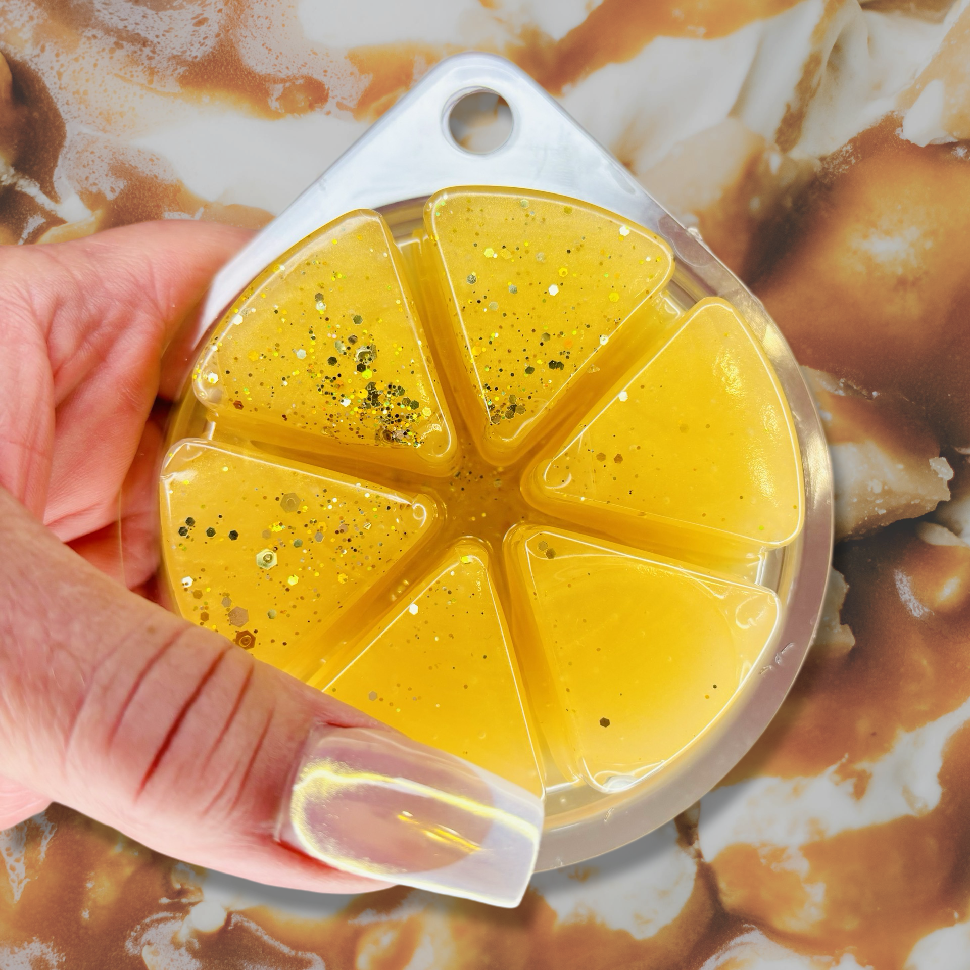A hand holds a round container featuring six yellow, glittery jelly segments of The Soap Gal x Fresh Churros Magical Gel Wax Melts 85g against a creamy, textured background. These luxurious, long-lasting fragrances envelop your space in an inviting aroma.