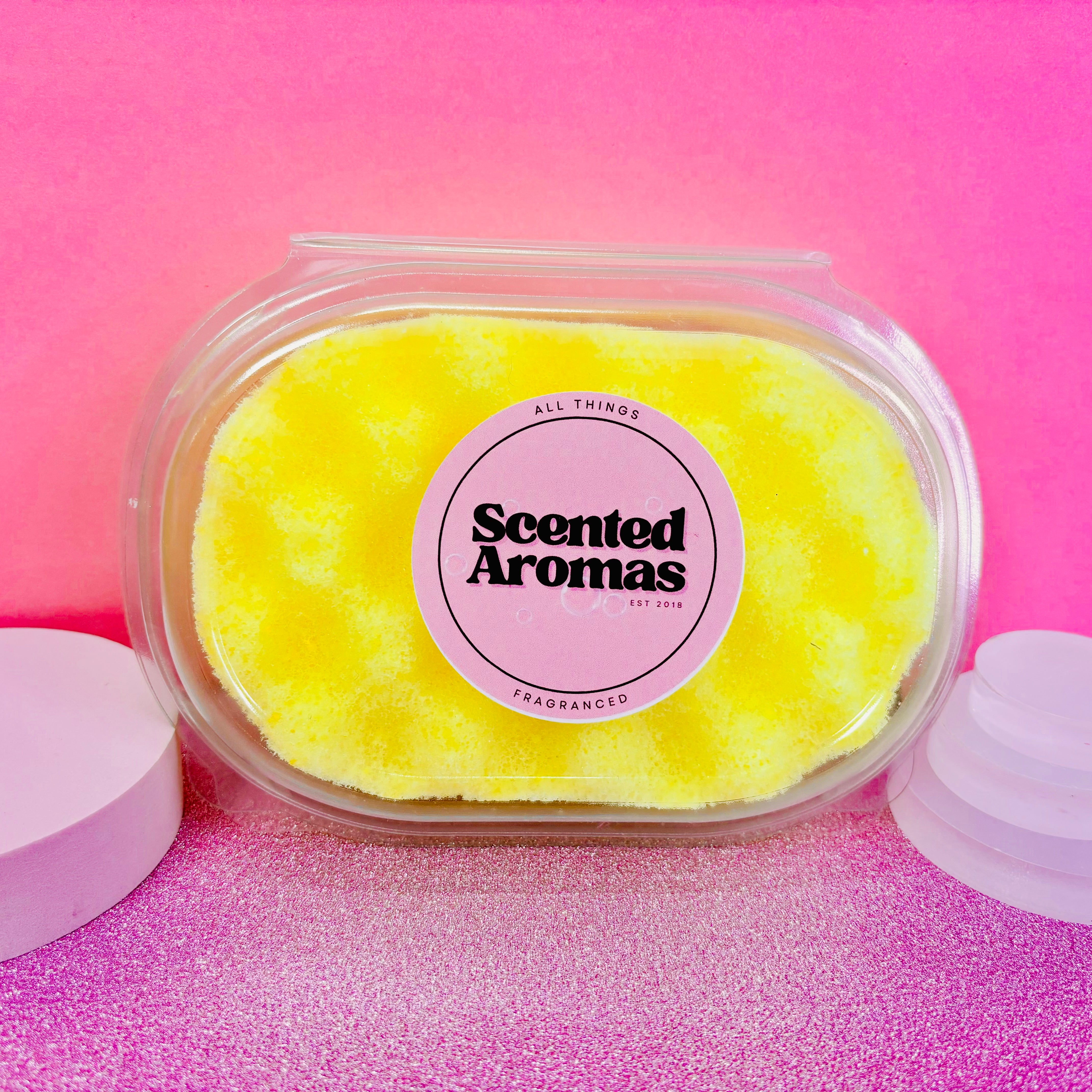 A container of yellow Citronella Soap Sponge by The Soap Gals, featuring natural insect repellent properties, is set against a pink background.