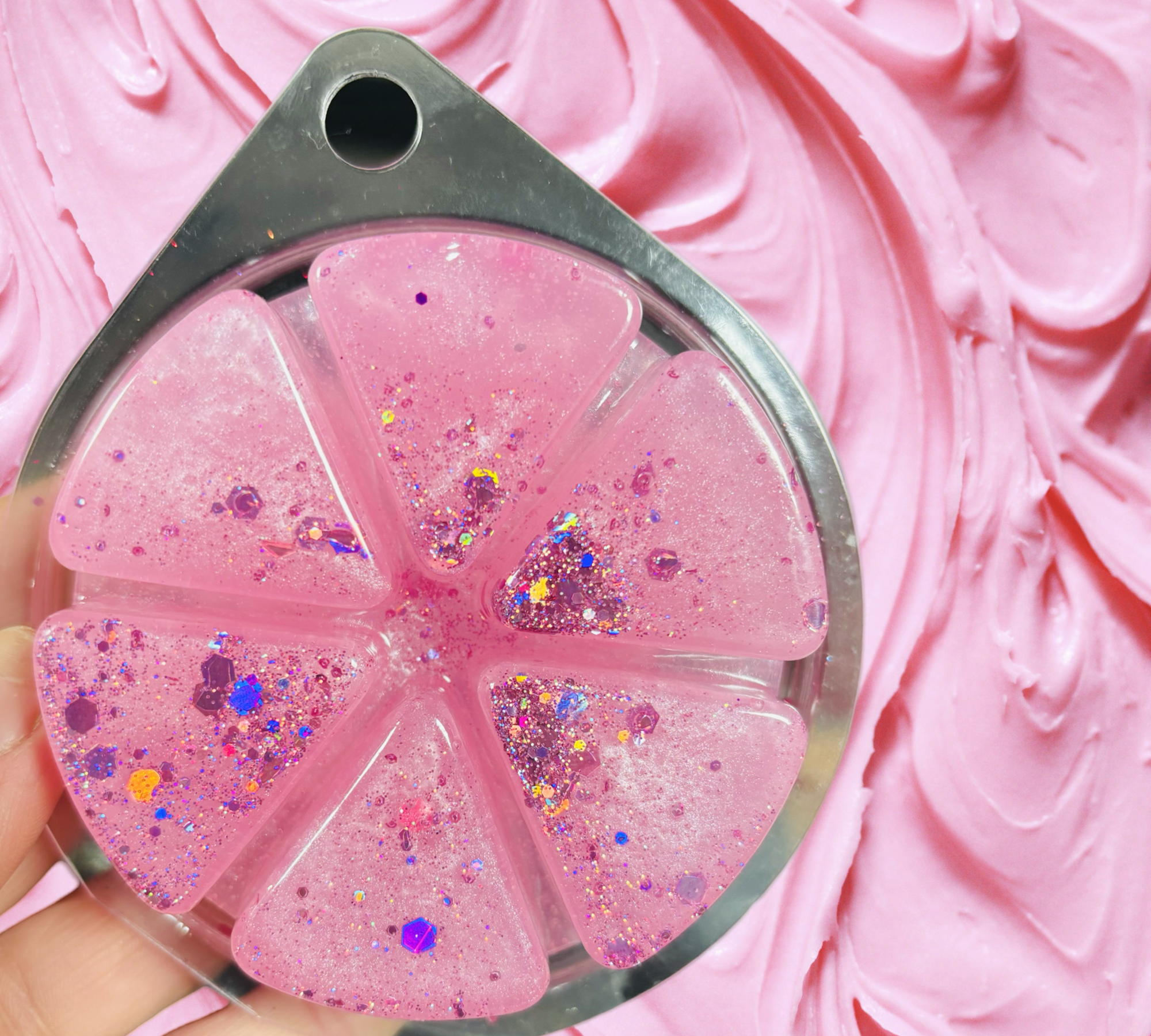 A hand holds a clear, pink, glittery citrus-shaped gel palette against a pink background—ideal for adding sparkle to the Mothers Day Gift Set Bundle by Scented Aromas, featuring five beauty and home fragrance products.