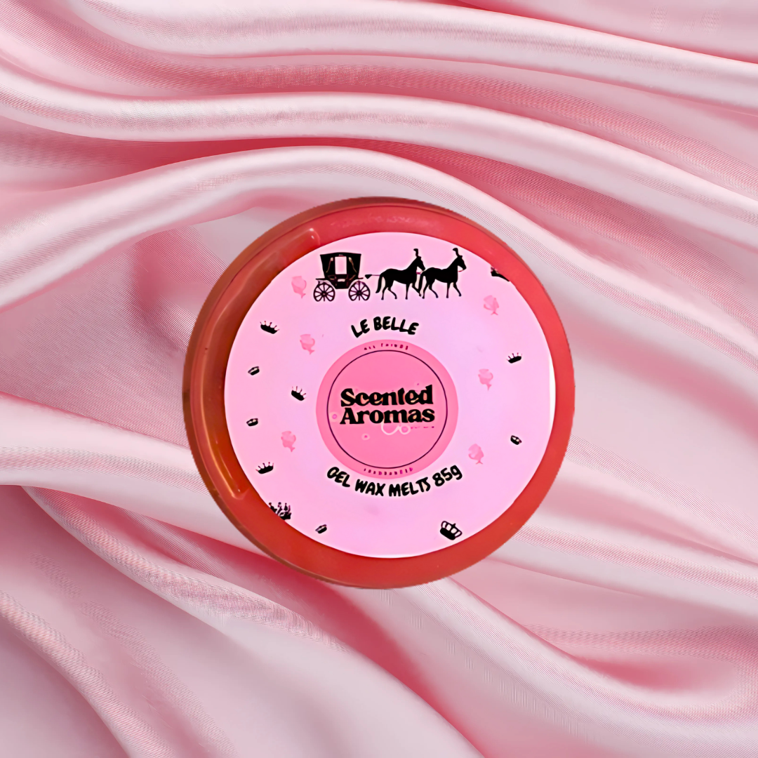 A round pink container of Le Est Belle Gel Wax Melts by The Soap Gal x rests on soft, pink satin fabric. The lid features a horse-drawn carriage design, hinting at the elegance within these luxurious perfume-scented wax melts.