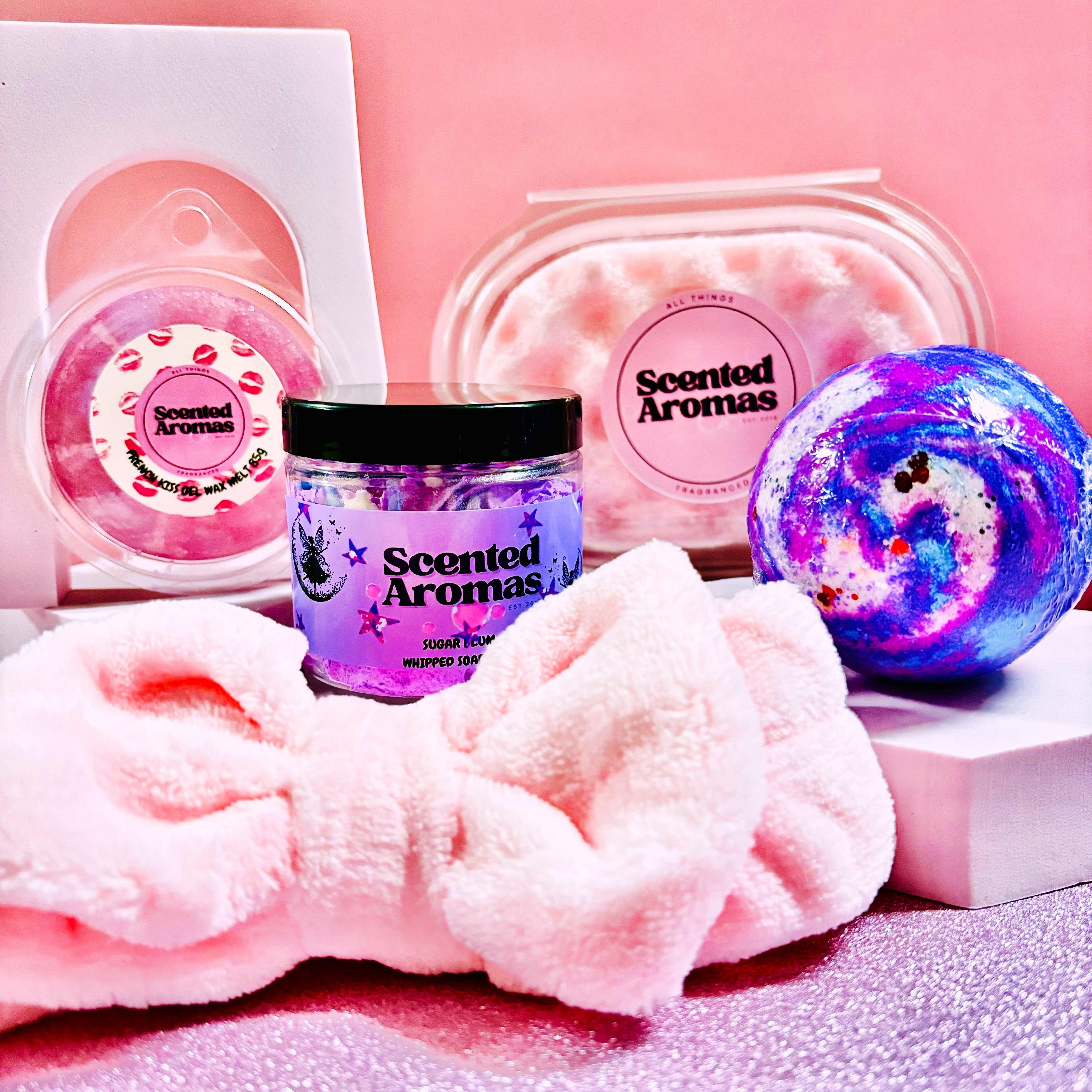 The Scented Aromas Mother's Day Gift Set Bundle includes a jar, two soap bars, a Birthday Cake bath bomb, and a pink headband, all beautifully displayed against a pink background. Perfect for celebrating Mother's Day!.