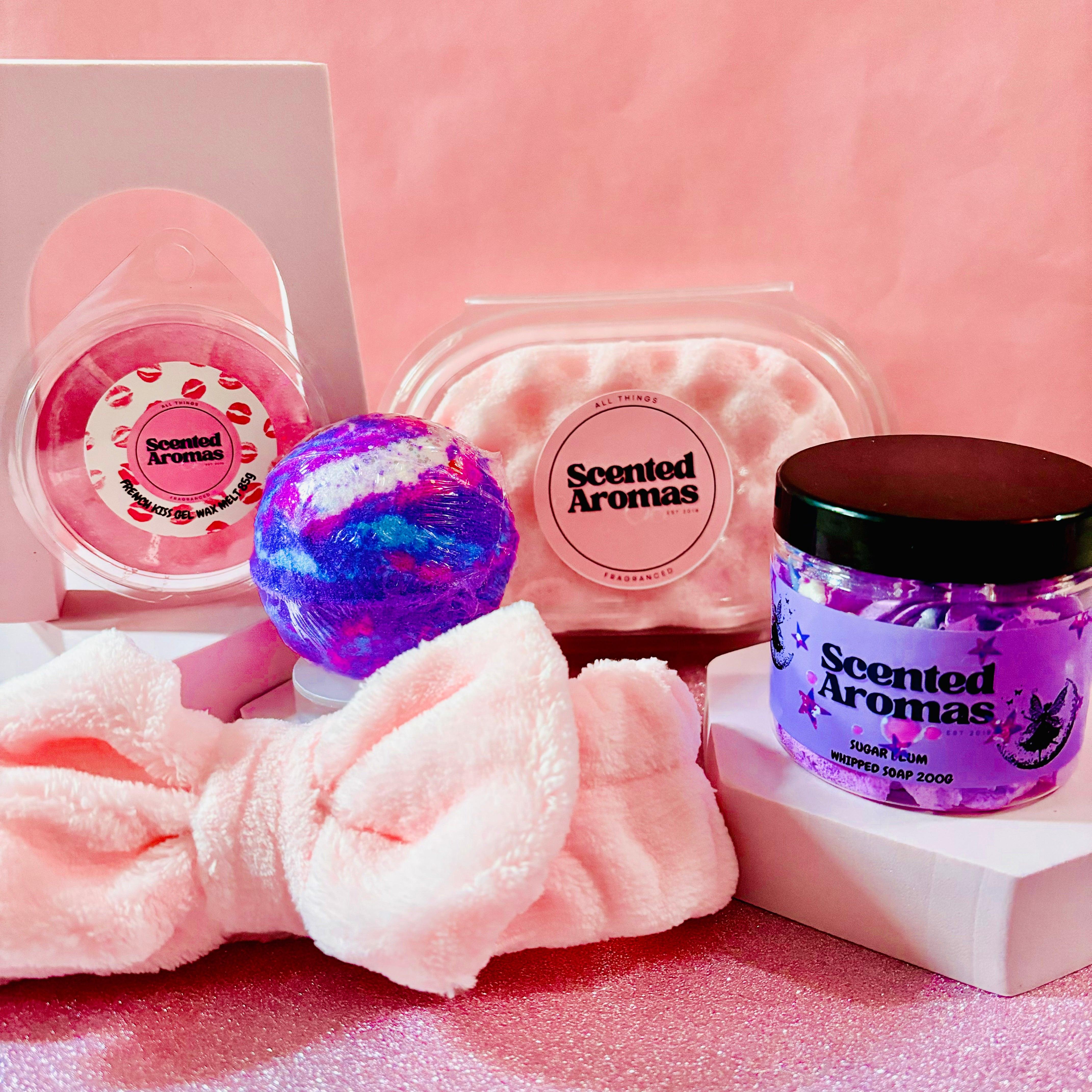 The Scented Aromas Mother's Day Gift Set Bundle offers five beauty and home fragrance products, including a Birthday Cake scented bath bomb, candle, jar, and pink hairband, arranged on a pink backdrop for soothing relaxation.