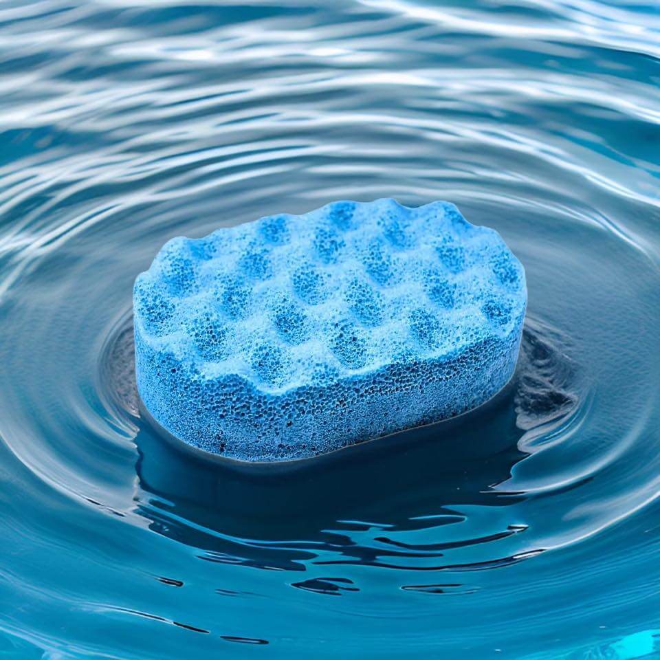 A blue, vegan sponge with a textured, honeycomb-like surface floats on calm, rippling water, reminiscent of the gentle touch of The Soap Gal x Peppermint Insect Repellent Soap Sponge.